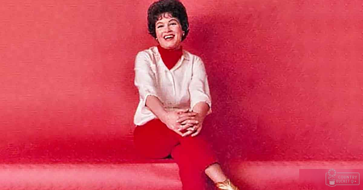 A middle-aged adult female in a relaxed fit white blouse with long sleeves and high collar, wearing gold high-heeled shoes against a solid red background.