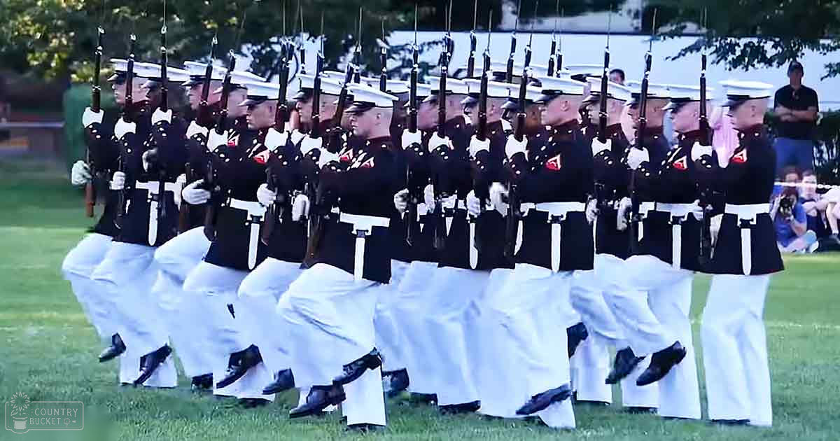 A group of young adult male soldiers in white and black uniforms with red shoulder patches stand in formation on a grassy field during an outdoor military parade.
