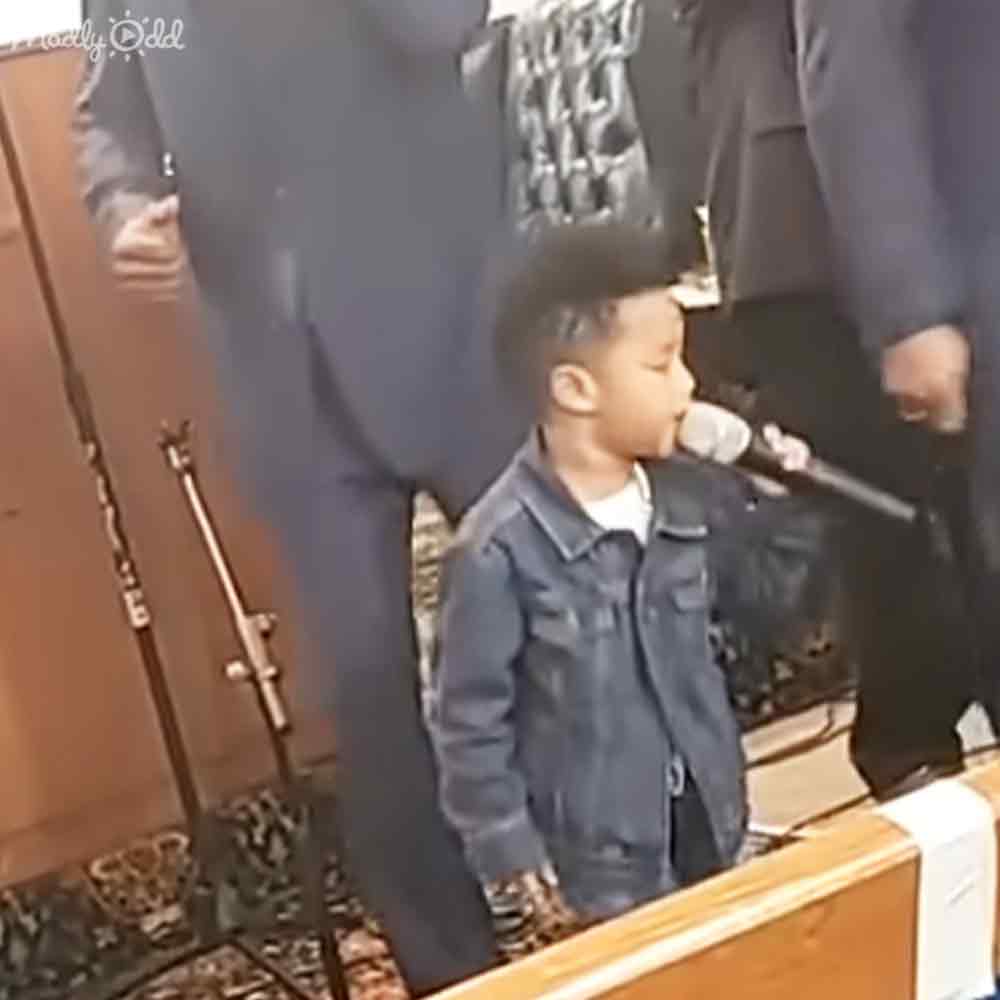 A young male singer wearing a dark denim jacket at an indoor event with a wooden podium and microphone stands in front, suggesting a performance or public speaking setting.