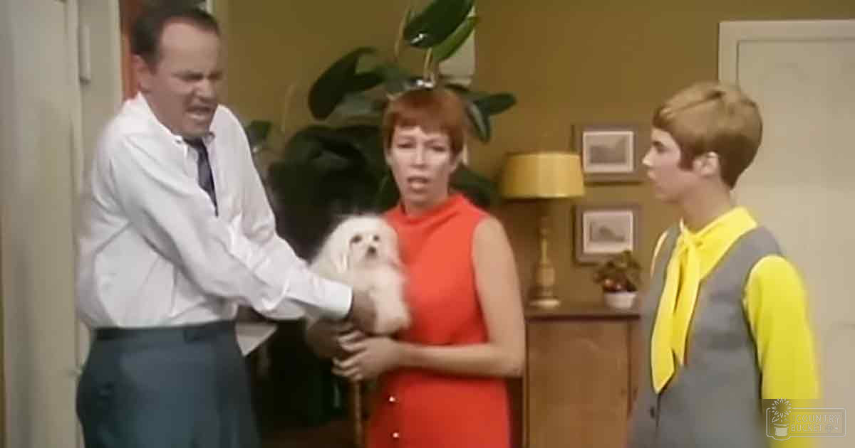 A family scene in an indoor setting with a middle-aged adult male, a possible middle-aged or older adult female, and a young adult boy. A small white dog, possibly a Maltese, is being held by the woman.