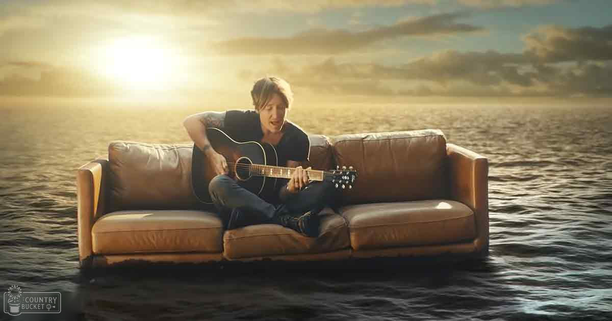 Young adult male playing guitar while sitting on a floating brown leather couch near the ocean, with partly cloudy skies and sunlight breaking through.