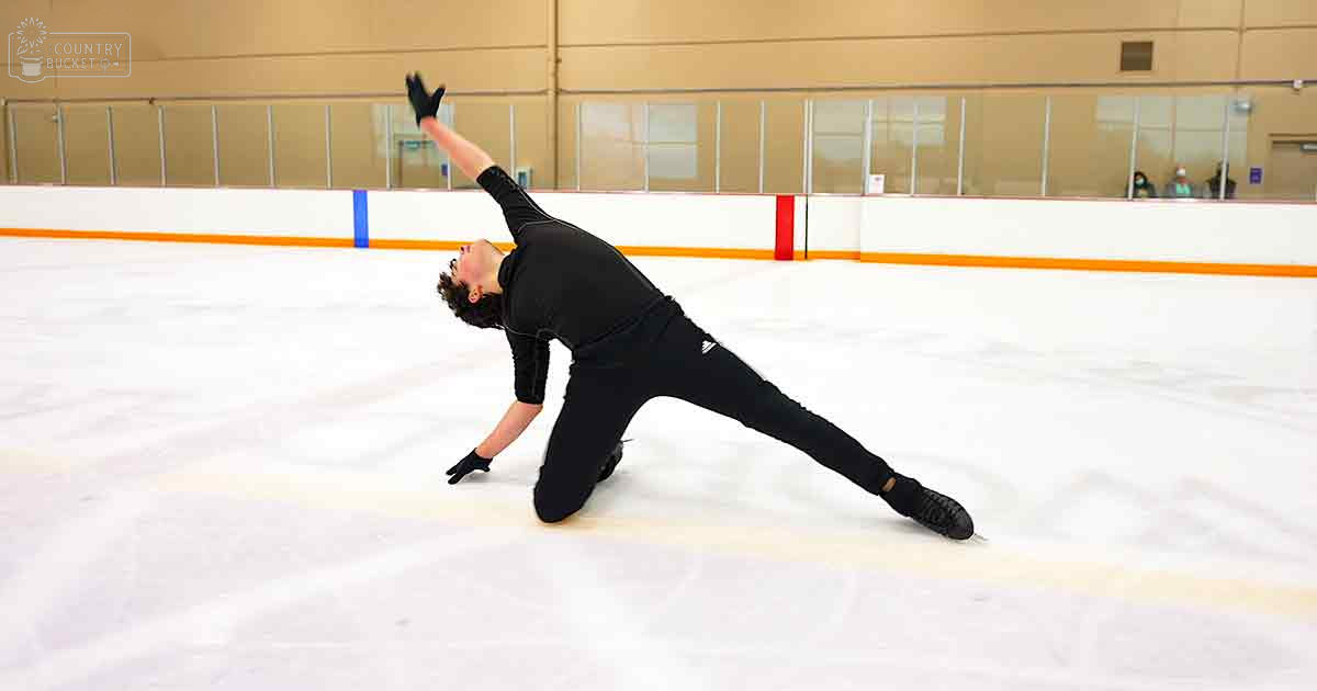 A young adult male figure skater in a black long-sleeved skating outfit stands on an indoor ice rink with white ice, orange barriers, and windows allowing natural light. Spectators are visible through the windows.