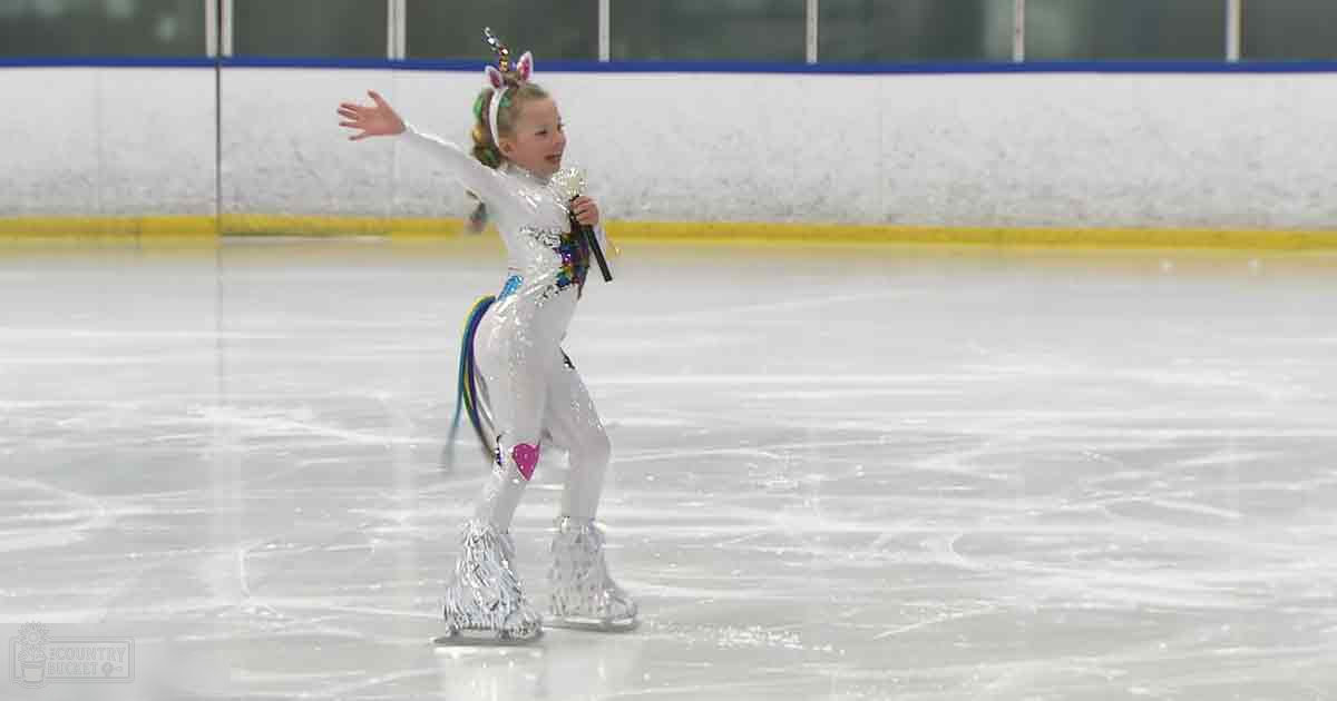 A child in a white leotard with colorful accents and ice skates at an indoor ice skating rink, well-lit daytime scene.