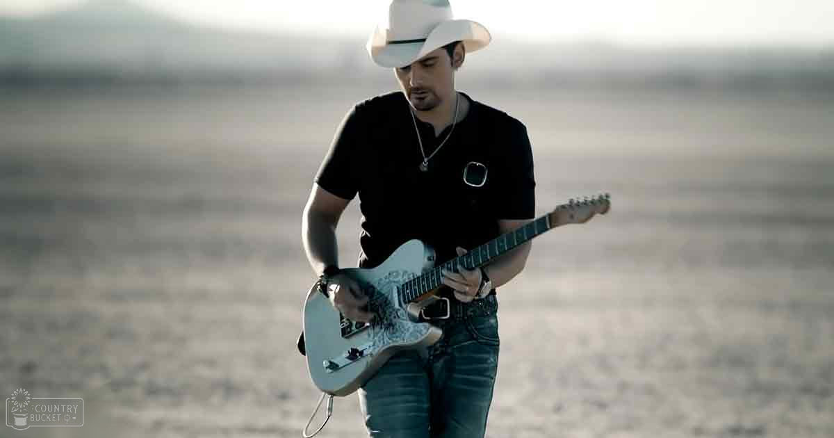 An adult male playing a white electric guitar with blue accents in an open, desert-like landscape under warm evening light.