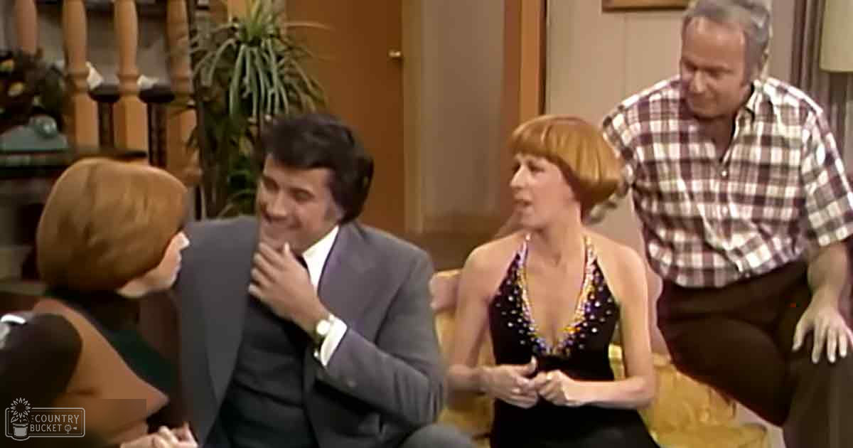 Indoor living room scene with a couch, staircase, potted plant, and lampshade; adult female in striped dress, middle-aged male in suit jacket holding hand near mouth, adult female in sequined dress, and middle-aged male in plaid shirt gesturing.