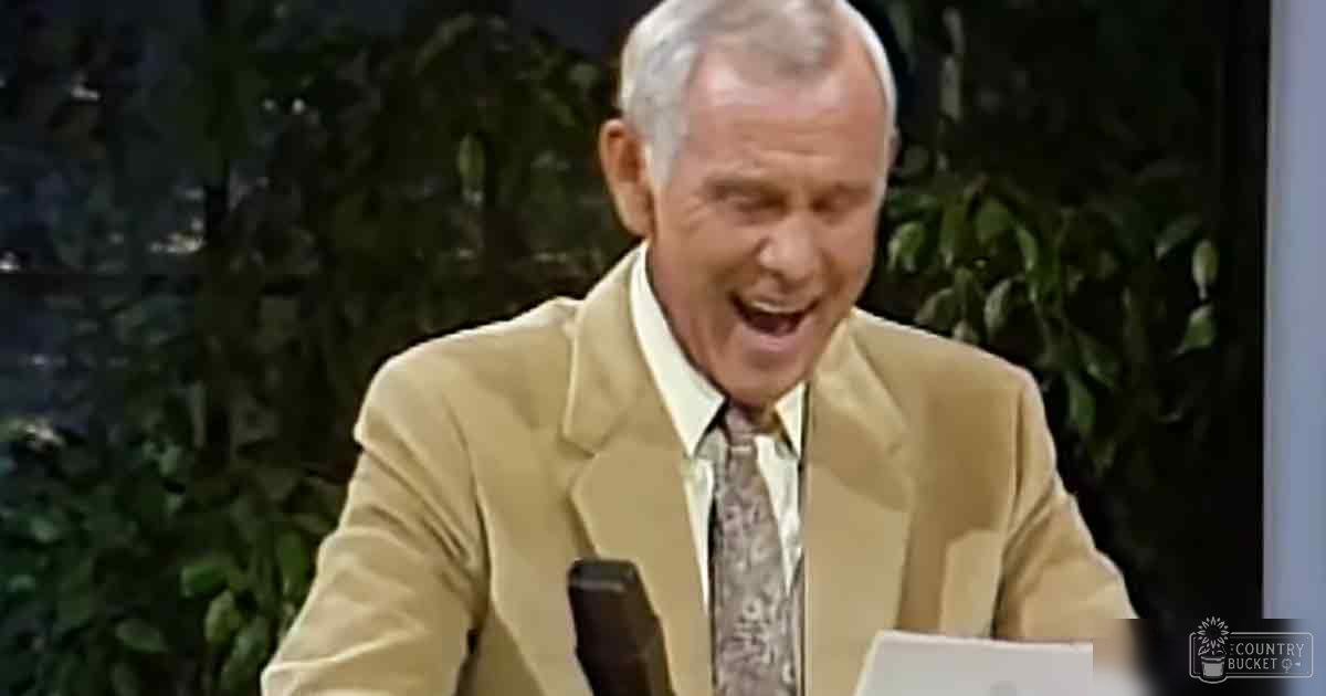 A middle-aged adult male wearing a light beige suit jacket with a patterned tie, seated indoors near a microphone on a desk. The setting suggests a professional or formal occasion in a studio environment.