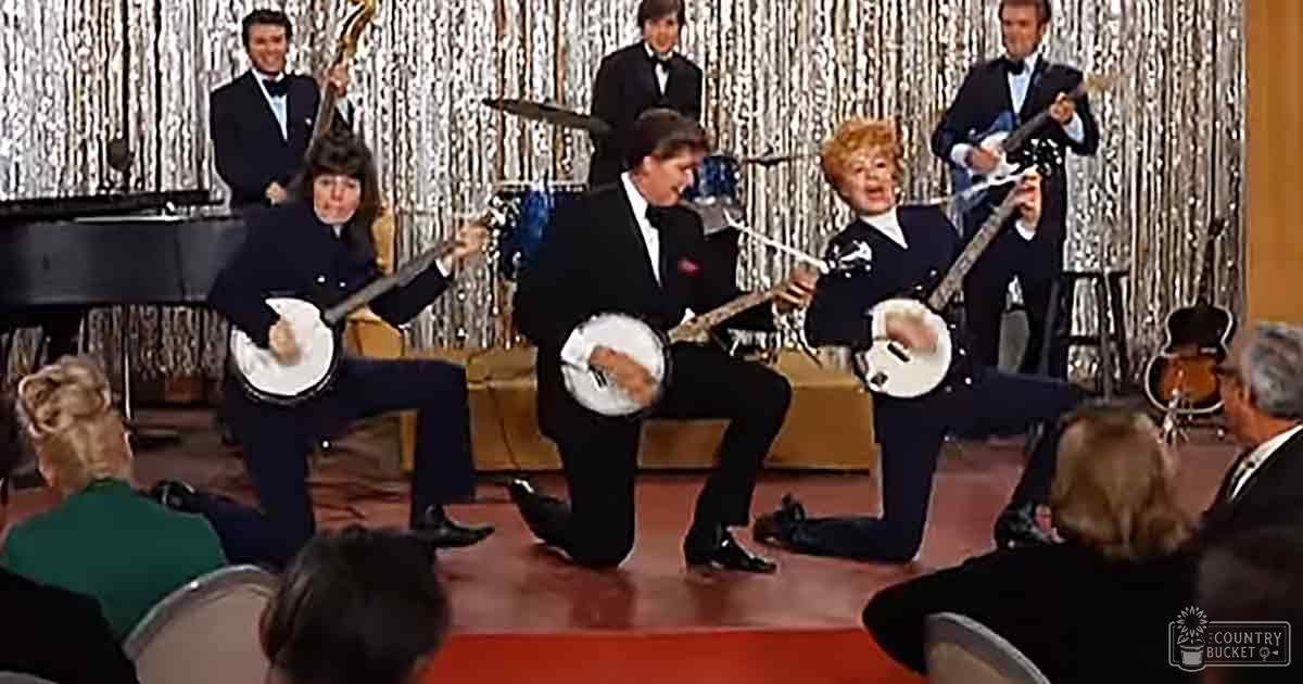 A musical performance scene with multiple adult males in dark suits playing banjos, a bass guitar, drums, and an electric guitar. A shiny reflective silver curtain is visible behind the stage.