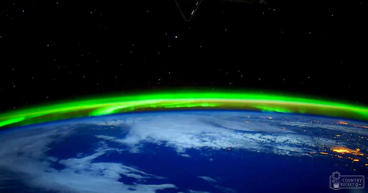 Aurora borealis over Earth's surface from space, nighttime scene with dark blue ocean, white clouds, green aurora, and black space backdrop.