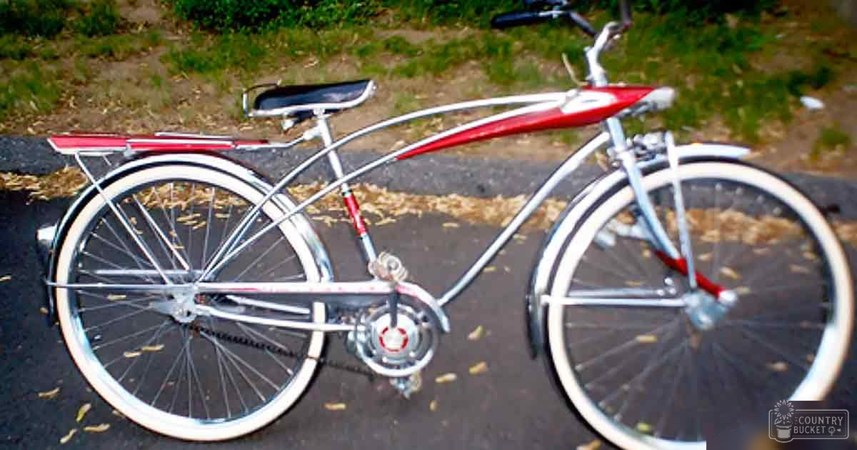 A vintage bicycle with chrome detailing, red accents on the frame, white-walled tires, black saddle and handlebars, small rear rack or fender attached to back wheel. Park-like setting with grassy areas, patches of dirt, fallen leaves indicating autumn daytime.