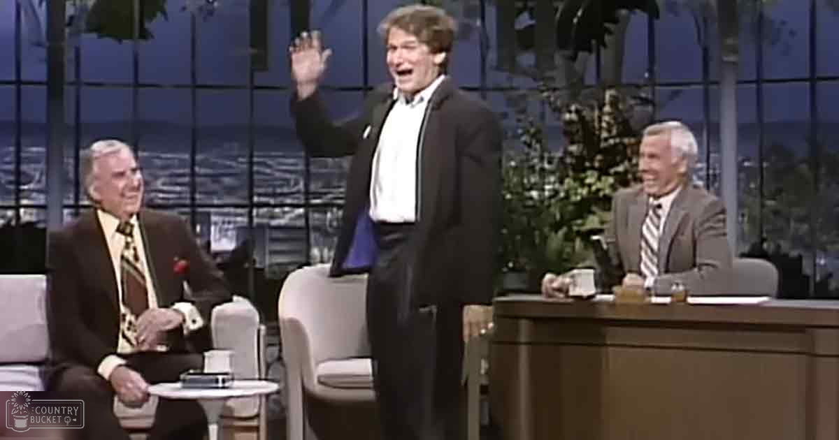 Indoor talk show setting with two older adult males and a young adult male in formal attire, large windows showing an urban night view.