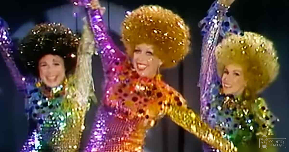 A performance scene with three adult females in glittery, sequined stage costumes with vibrant colors like purple, green, blue, gold, orange, red, black, and white under dim stage lighting.