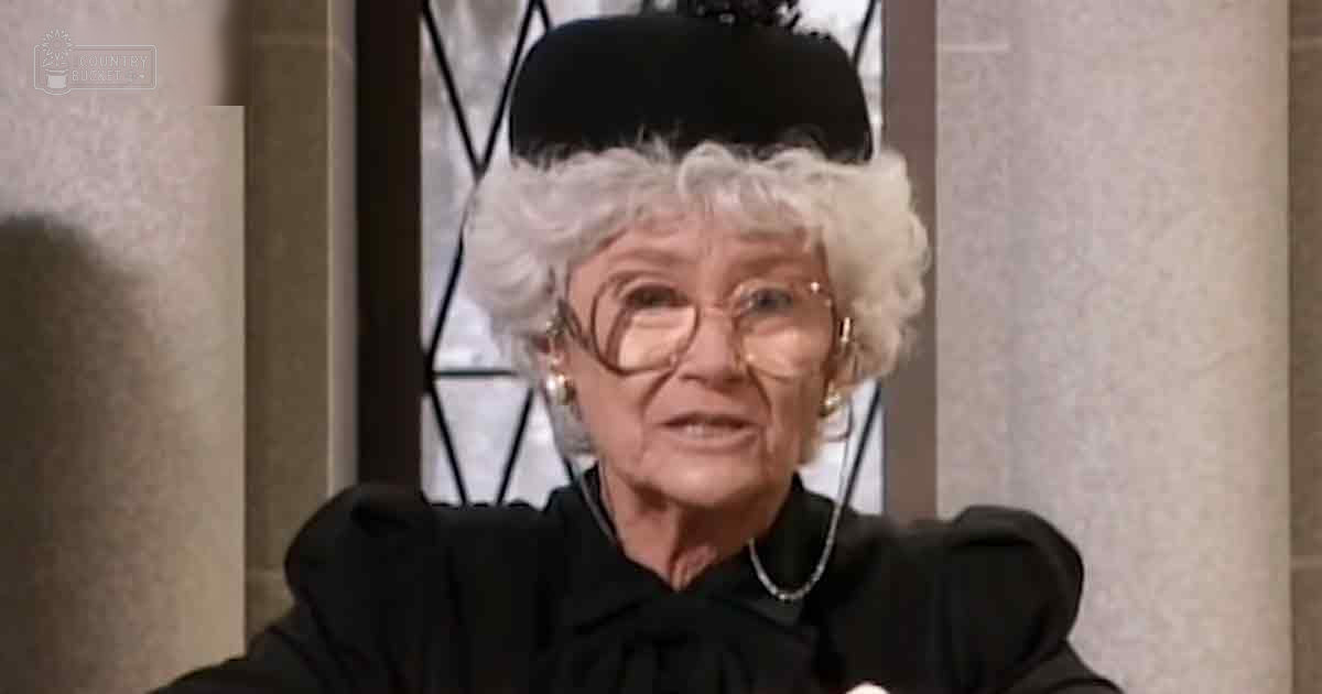 An older adult woman wearing a black, high-collared garment with puffed sleeves and large round glasses stands indoors near a window with decorative metal bars.