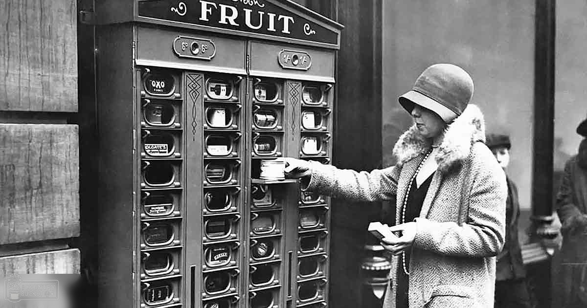 A woman in a fur-trimmed coat interacts with an early vending machine labeled 'FRUIT' in what appears to be an urban public setting, possibly from the early 20th century.