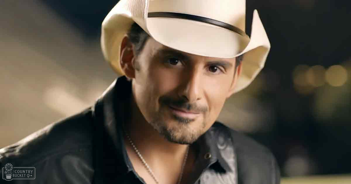 A middle-aged adult male in a fitted black button-up shirt with paisley pattern, wearing a white cowboy hat and silver necklace, stands indoors under warm evening lighting.