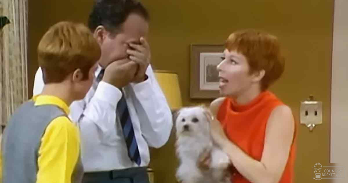 A young child in a yellow and gray shirt interacts with an adult male wearing a white dress shirt and blue striped tie, while an adult female in an orange top observes nearby. A small dog is visible in the background within a cozy indoor living room setting.