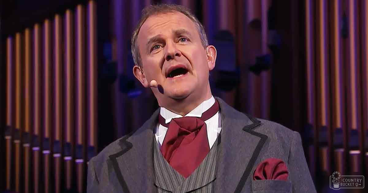 A middle-aged adult male in a formal gray suit with white shirt and red bow tie performing on stage, with an organ backdrop.