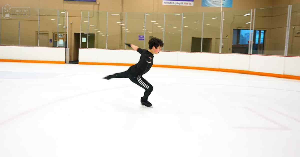A young adult male figure skating in an indoor ice rink with glass walls, orange barriers, and signage reading 'watch it, play it, live it'.