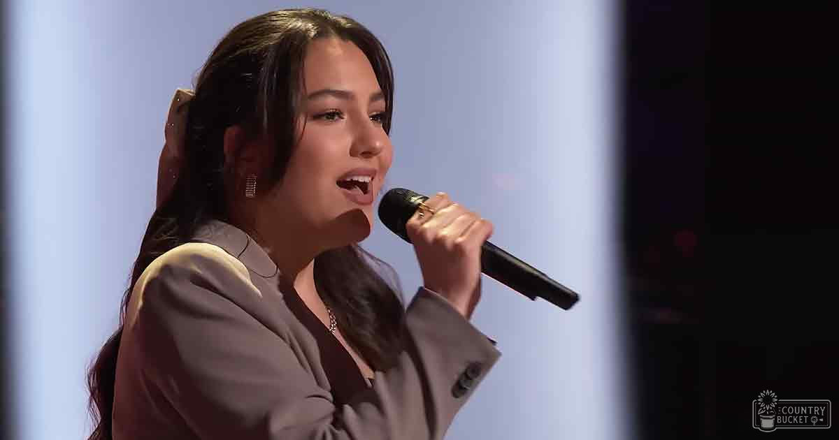 A young adult female in a light brown blazer sings into a microphone against stage lighting, suggesting an indoor performance setting.