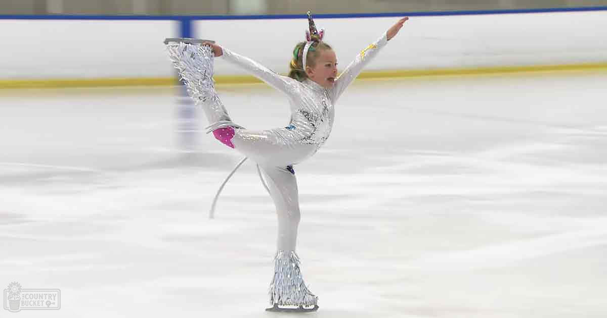 A young female figure skater in a shimmering silver and pink figure skating costume with long sleeves, standing at an indoor ice rink.