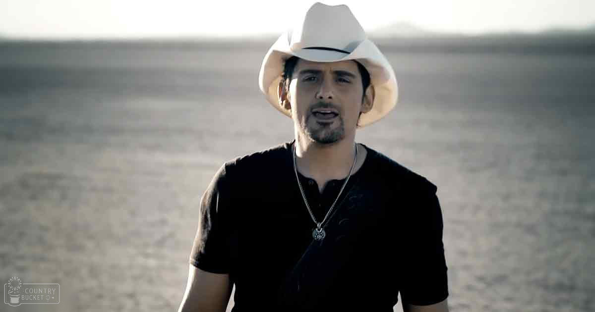 Adult male wearing a black, modern casual button-up shirt with relaxed fit, collared neckline, short sleeves, white cowboy hat, necklace with pendant, in an arid desert landscape during late afternoon daylight with warm tones.