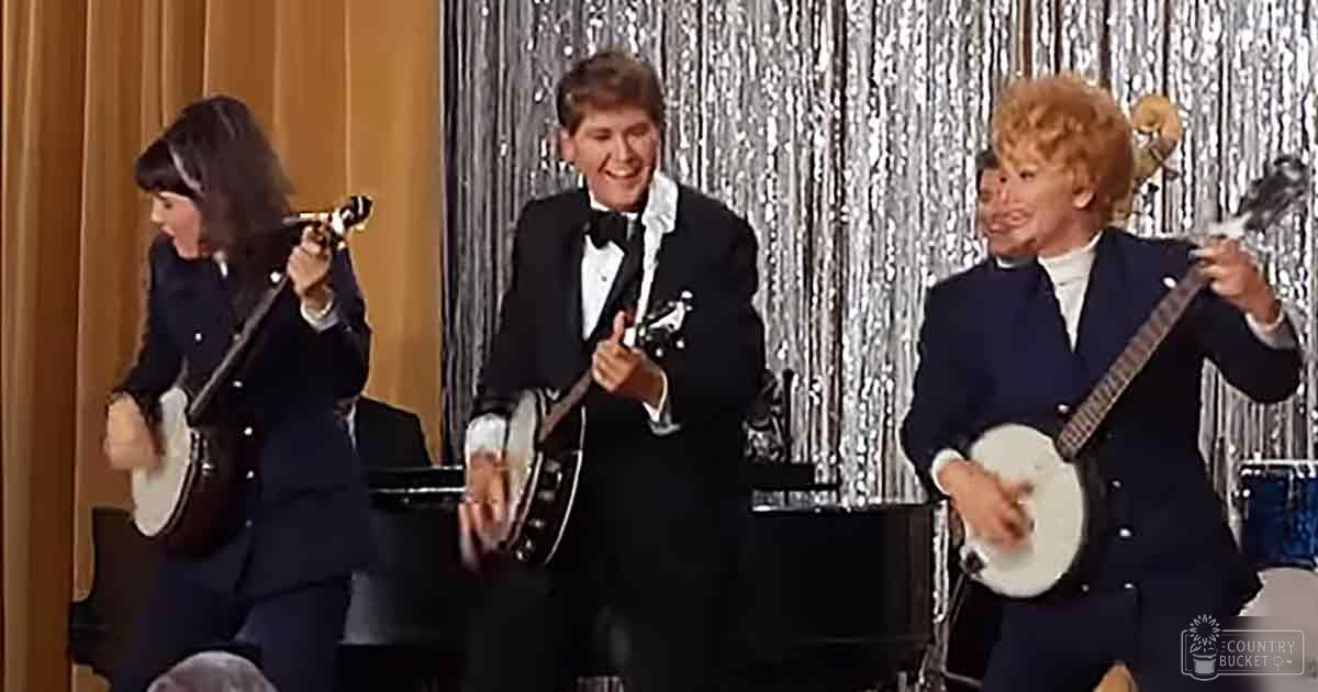 A young adult female in a dark suit with a white shirt and dark tie stands beside another young adult male in a tuxedo with a black bow tie, both part of a mid-20th century musical performance set in a studio environment.