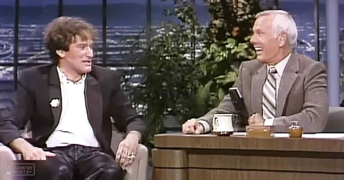 Two adults in a talk show setting, one wearing a dark blazer and the other in a light gray suit, with cityscape background and evening lighting.