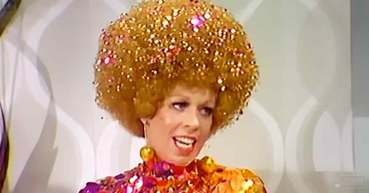 A female performer in a theatrical setting, wearing a bright pink and gold sequined top with high neckline and short sleeves. The backdrop is white with circular patterns.