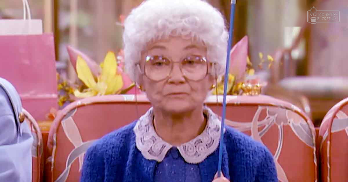 An older adult female wearing a blue knitted sweater with lace trim, large-rimmed glasses, in an indoor home setting with vintage furniture and warm tones.