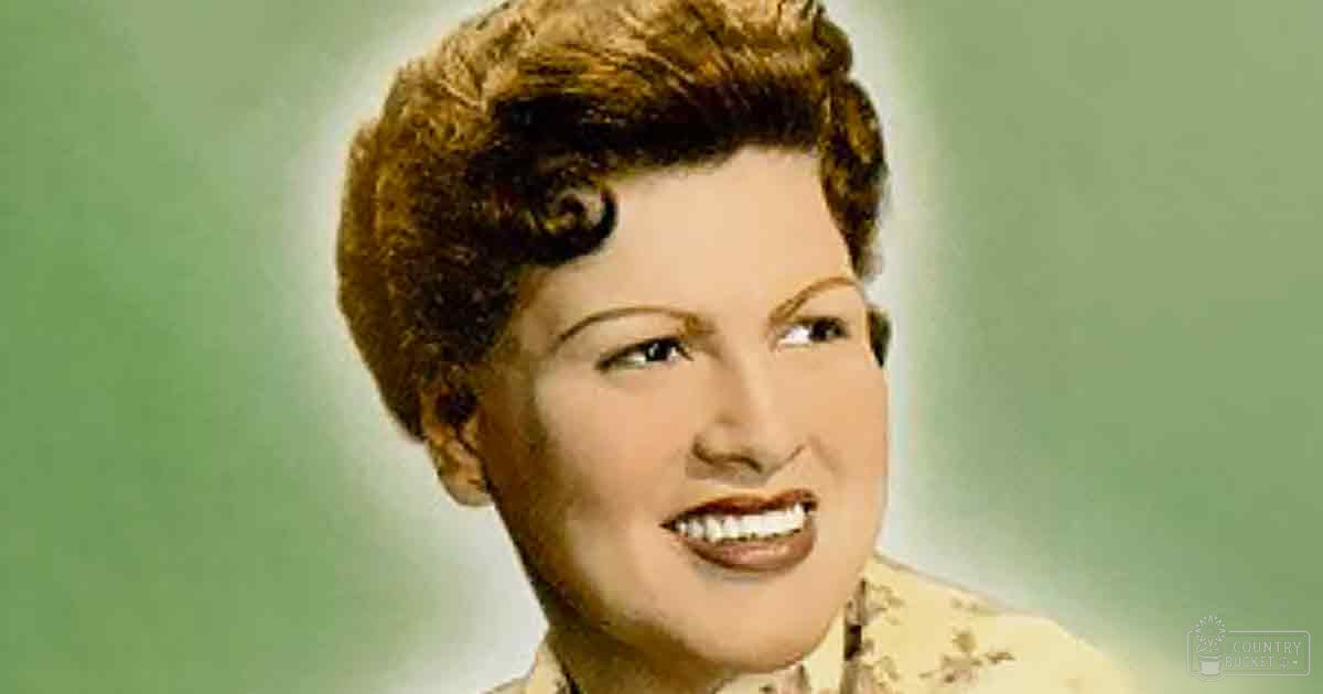 A middle-aged adult female stands against a plain light-green indoor backdrop wearing a relaxed, collared, long-sleeved, beige or light tan floral-patterned blouse with brownish-red accents and a cotton or polyester blend fabric. The setting appears to be designed for portrait photography under even artificial lighting.