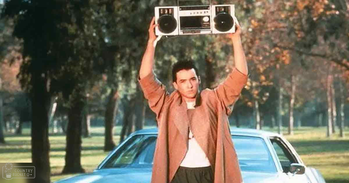 A young adult male in a long brown coat stands outdoors, holding an old-style boombox above his head with both hands. Behind him is a classic car from the 1970s or early 1980s.