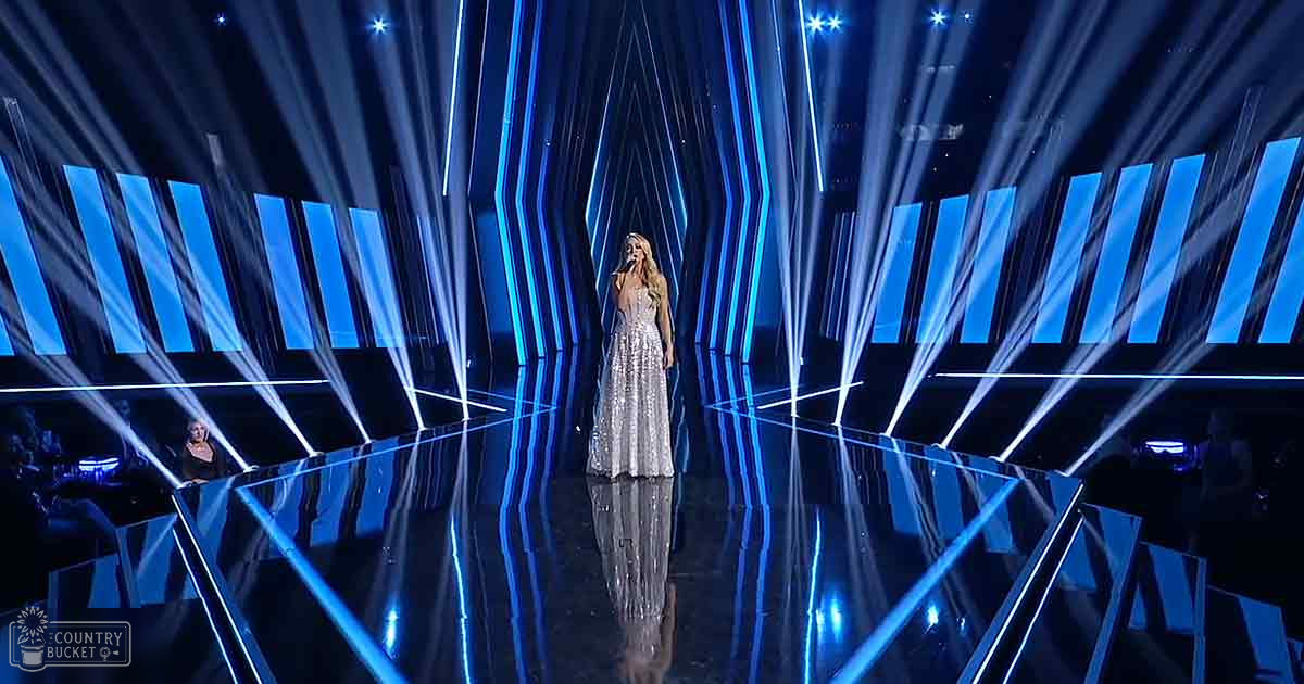 A young adult female performer in a shimmering silver or white sequined gown with a deep V-neck on stage under bright blue lights.