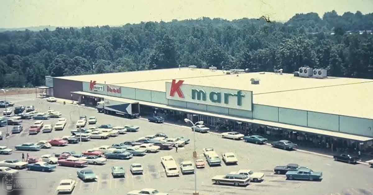 Kmart store outdoors with parked 1960s or early 70s era cars, trees in background, summer setting, daytime light