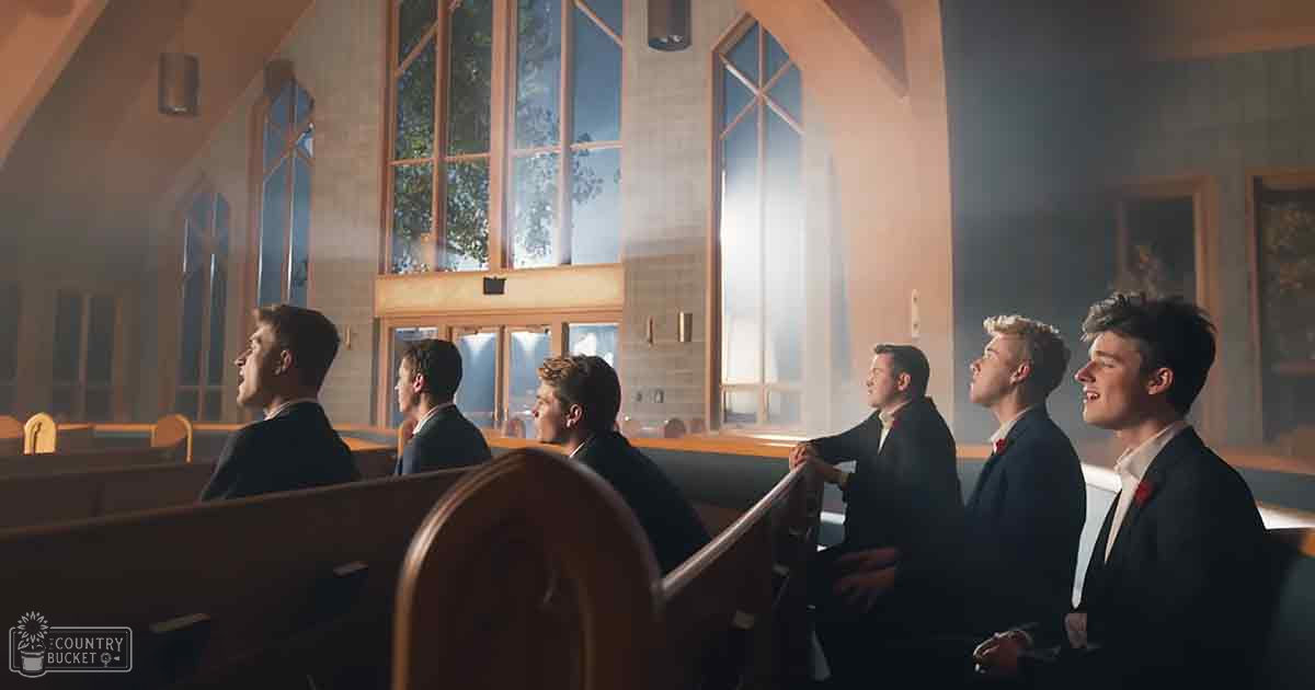 Young adult males in dark suits seated on wooden pews inside a church with large stained glass windows and natural light streaming in, suggesting an indoor daytime or early evening setting.
