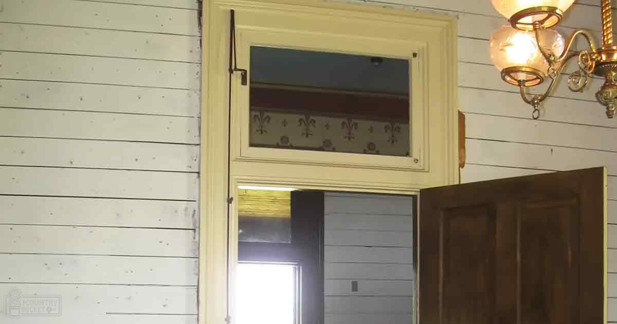 An indoor scene showing a partially open wooden door, white-painted walls with horizontal planks, an upper frosted glass window with decorative pattern, and a chandelier emitting warm light. The setting appears to be a room in a house or similar structure.