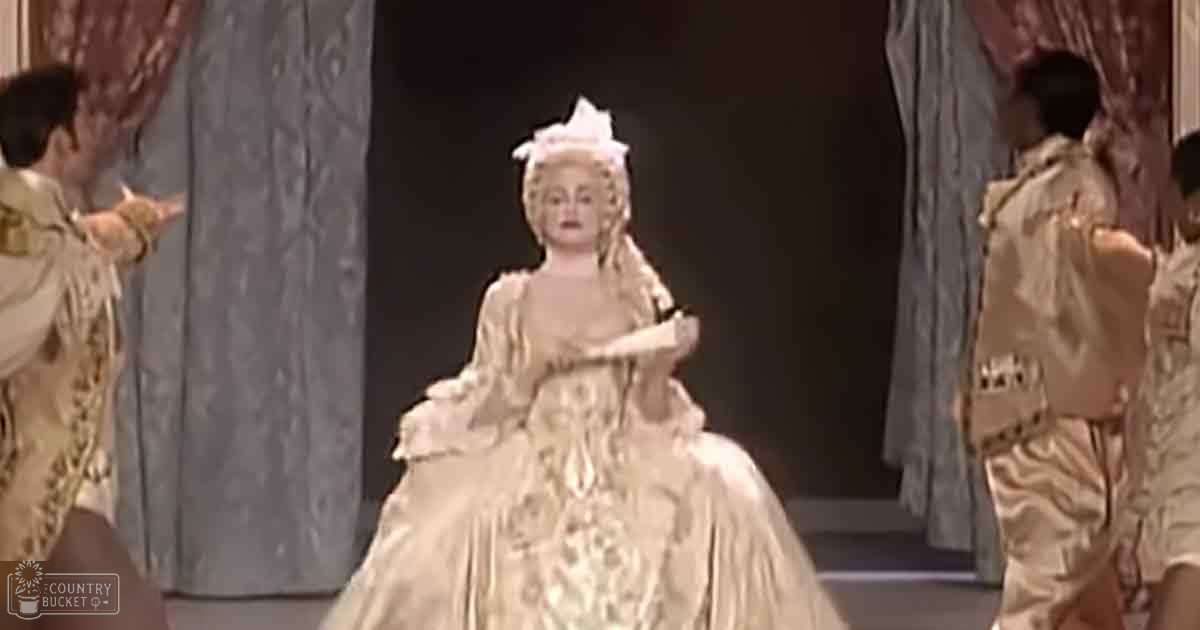 A woman in an elaborate, light-colored 18th-century gown stands on a stage with other individuals in period attire. The setting is indoors and appears to be theatrical.