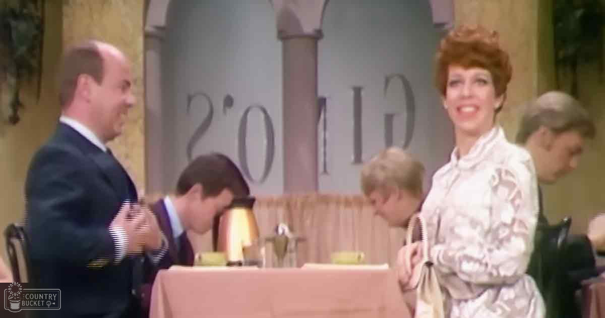 An adult female in a restaurant wearing a light-colored, patterned blouse with long sleeves over a dark suit jacket; a child sits beside her at a table with a lamp and purse.