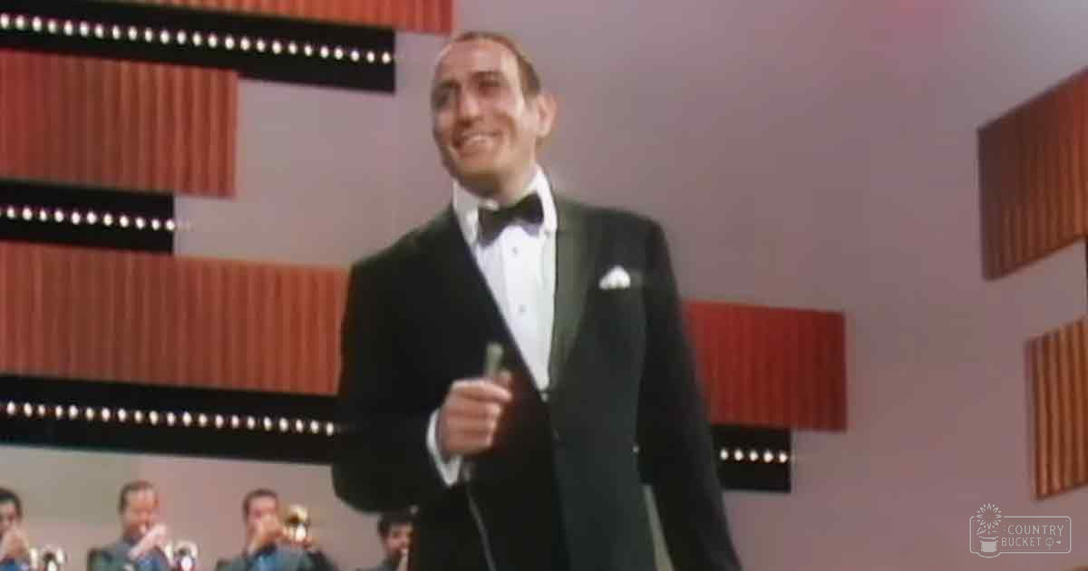 An adult male singer in a well-fitted tuxedo with white shirt, black bow tie, and white pocket square performing on stage at an event or performance venue.