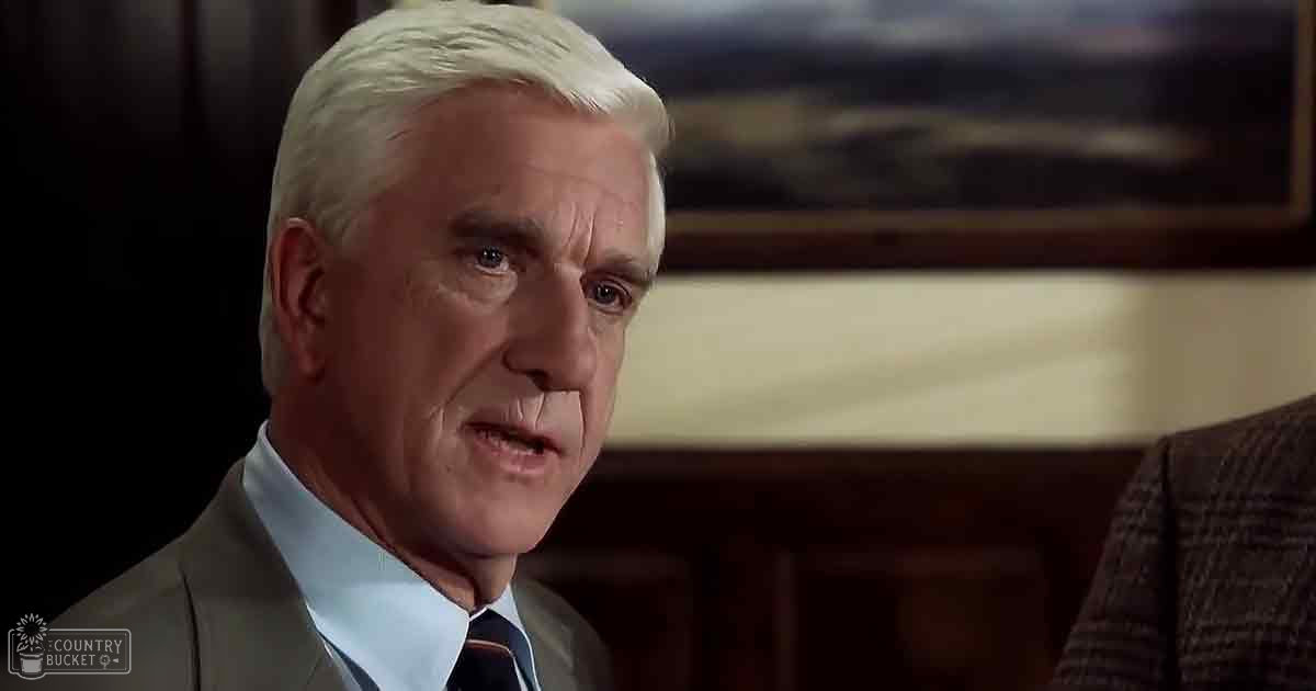 An older adult male in a formal setting, wearing a light gray suit jacket, white dress shirt, and striped tie, indoors with dark wood paneling and framed artwork.