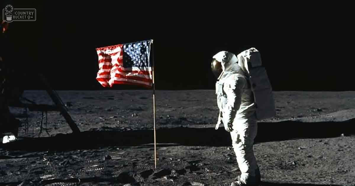 An astronaut in a full spacesuit stands near an American flag on the Moon's surface at night.