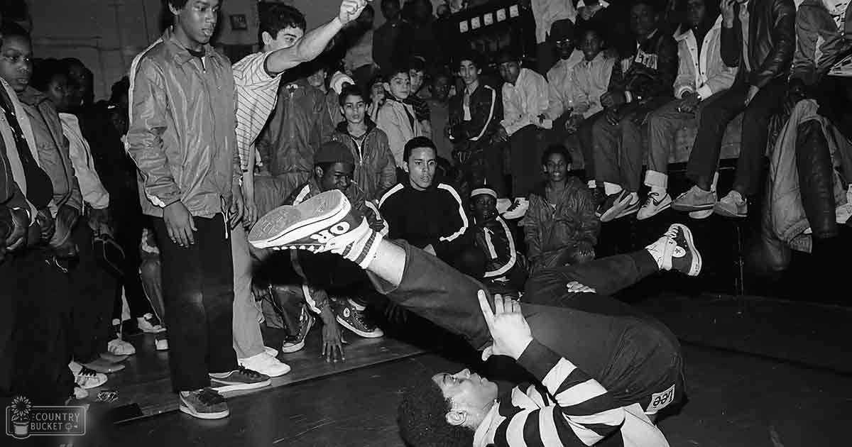 A young adult male breakdancer performs a handstand on an indoor wooden floor while spectators watch from the sides and background, dressed in casual late 20th-century streetwear.