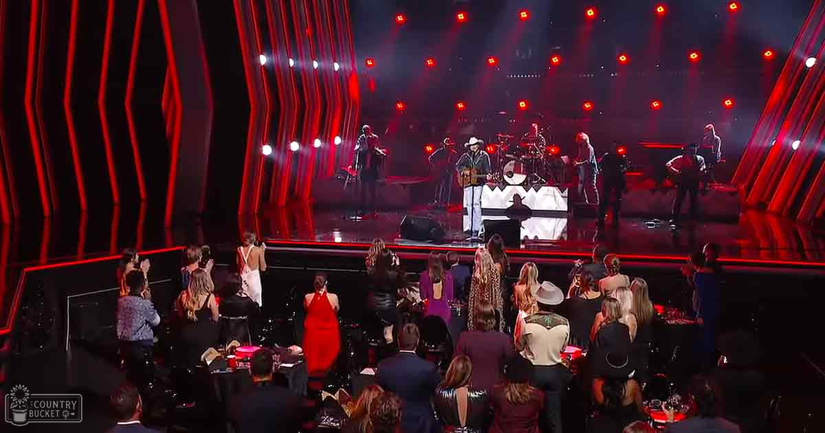 Live music performance scene at indoor concert venue, audience dressed in formal and casual attire under artificial evening light, red and black dominant colors with a large screen displaying the stage.