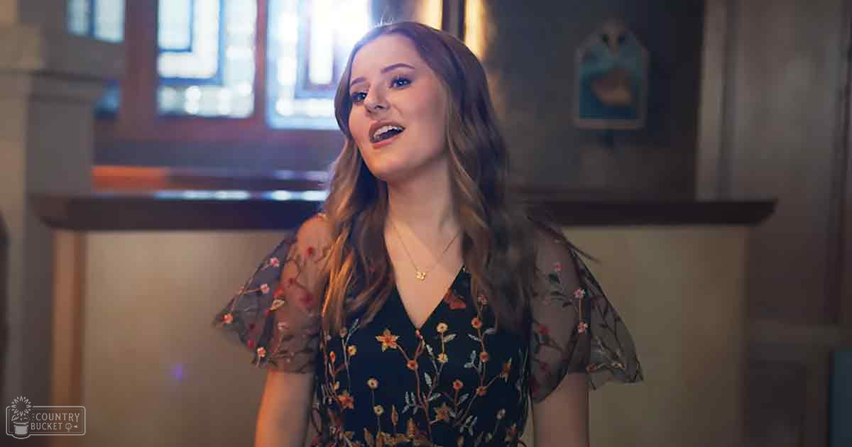 A young adult female wearing a dark floral dress with puffed sleeves and gold necklace, indoors in a home setting.