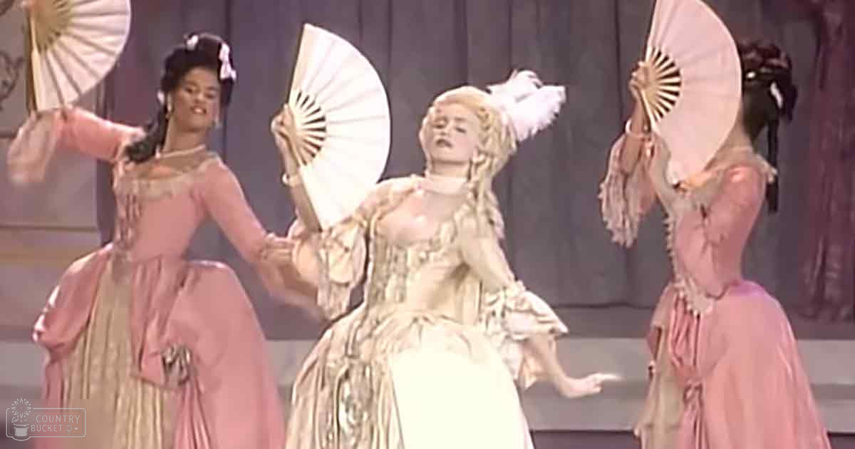 Three adult female performers wearing ornate gowns with puffed sleeves and high necklines, possibly from an opera or historical drama production, holding fans against a stage backdrop.