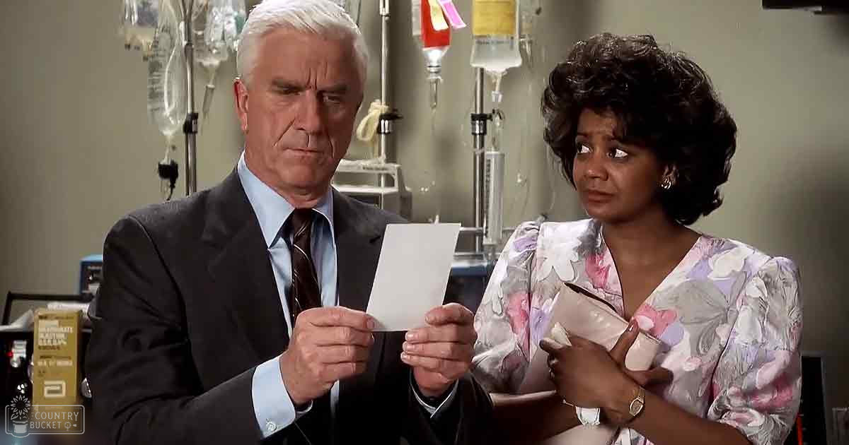 An office or medical environment scene with two adults, one male and one female. The male wears a dark gray suit jacket over a light blue dress shirt and dark tie, while the female dons a floral blouse with long sleeves and a V-neckline. Medical equipment is visible in the background.