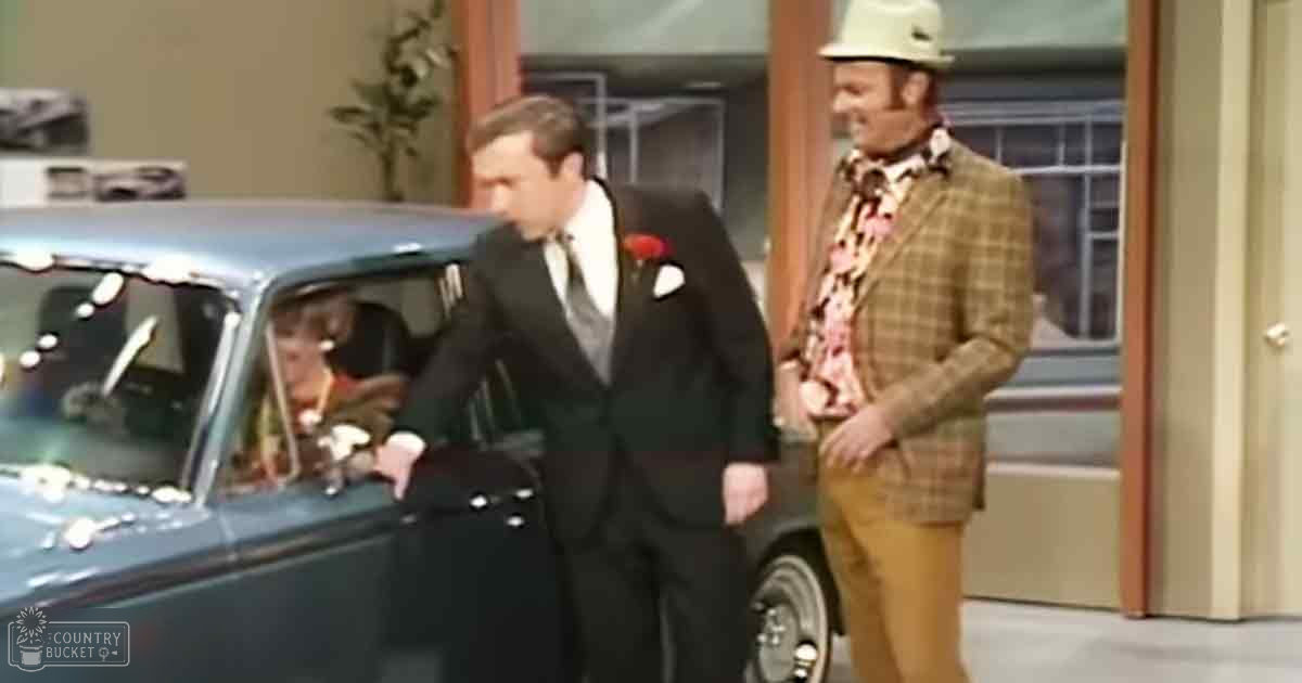 Adult male in a tuxedo with a red boutonniere stands indoors at what appears to be a formal event like a wedding; another adult male wears a plaid blazer and beige fedora, while a child in light clothing is seated inside a classic car from the 1960s or early 1970s.