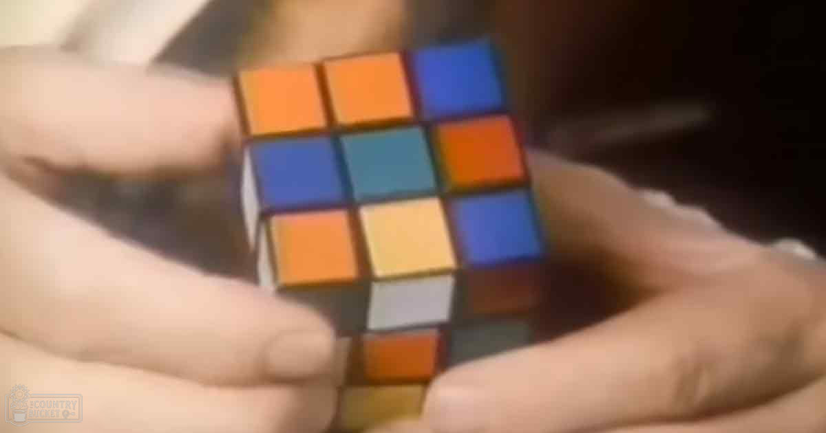 An adult male holds a Rubik's Cube with 'Madly Odd' text in the bottom left corner, indoors setting with blurred background.
