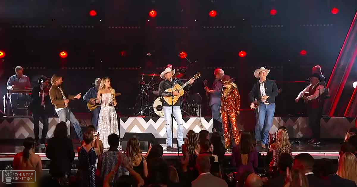 Musical performance in an indoor arena with red spotlights, evening lighting, and diverse audience attire.