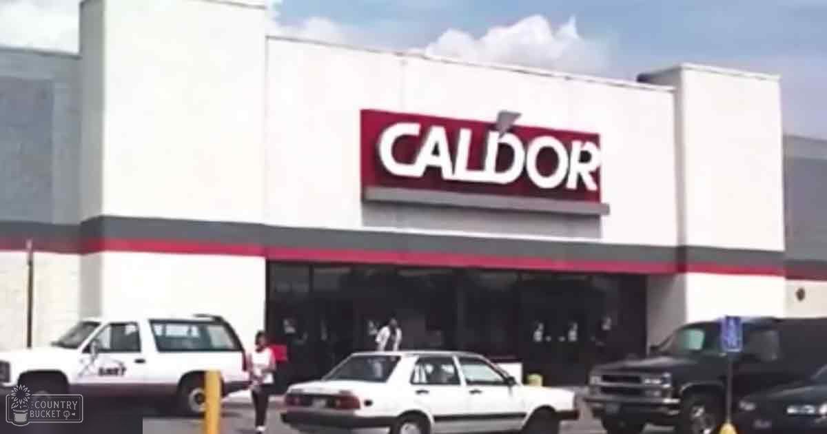A Caldor store in an urban area with two individuals; one young adult female wearing a white t-shirt, dark pants, and carrying something; another older adult male partially obscured by vehicles on the left. Vehicles include a white SUV labeled 'LMT', a white sedan, part of another vehicle.