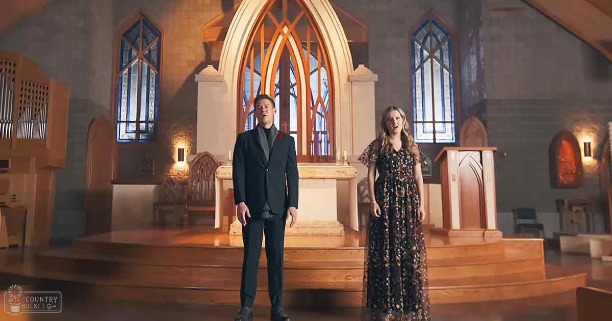 A man in a dark suit with a red rose singing alongside a woman in a floral dress at an altar inside a church, surrounded by stained glass and wooden elements.