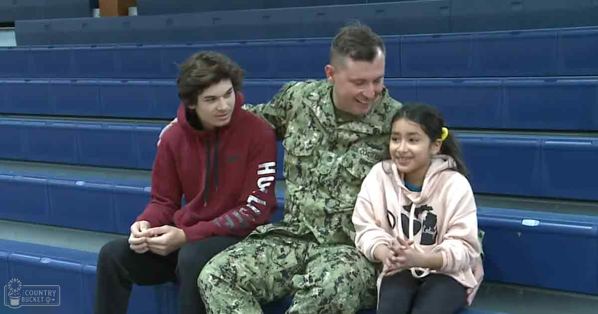 Children with an adult male in a military uniform at an indoor sports facility, likely gymnasium setting, daytime artificial lighting, blue bleachers, camouflage uniform, maroon hoodie, light pink hoodie, various clothing shades.